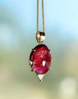 A gold chain necklace with a large, oval-shaped, deep red gemstone pendant. The pendant has a faceted design and is set in a gold setting.
