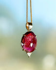 A gold chain necklace with a large, oval-shaped, deep red gemstone pendant. The pendant has a faceted design and is set in a gold setting.