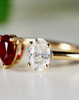 Moissanite & Ruby Engagement Ring, A gold ring with two oval-shaped gemstones. The larger gemstone is clear and the smaller gemstone is red.