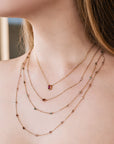 Four delicate gold necklaces of varying lengths. The top necklace features a small, oval-shaped ruby pendant. The other necklaces are adorned with small, colorful gemstones. The necklaces are layered, creating a sophisticated and elegant look.