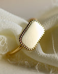 A gold ring with a rectangular, smooth, blank surface surrounded by a beaded border.