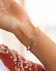 Gold-toned bracelets with delicate designs. One bracelet features a bar design, while others have small, colorful gemstones. The bracelets are worn on a wrist, showcasing their elegant and minimalist style.