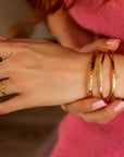 A woman wearing three hammered gold cuff bracelets in varying thicknesses, stacked on her wrist. Gold filled bangles with a personalized engraved message option.
