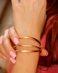 A woman wearing three hammered gold cuff bracelets in varying thicknesses, stacked on her wrist. Gold filled bangles with a personalized engraved message option.