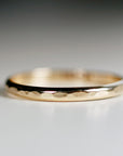 Close-up of a delicate gold hammered wedding band. The band features a subtly textured, hammered surface, adding a touch of rustic elegance. Its simple design and warm gold hue make it a timeless and versatile choice for everyday wear. The band&
