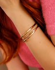 A woman wearing three hammered gold cuff bracelets in varying thicknesses, stacked on her wrist. Gold filled bangles with a personalized engraved message option.