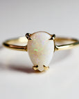 A gold ring featuring a pear-shaped opal gemstone. The opal has a milky white base with iridescent flashes of color. The ring&
