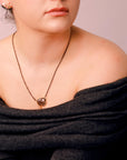 A woman wearing a black off-the-shoulder sweater with a delicate chain necklace featuring a brown gemstone pendant.