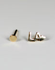 A pair of gold earrings. One earring is a small circle, the other earring says "LA" with a heart.