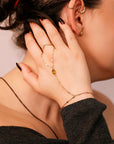 A gold hand chain bracelet with a teardrop-shaped citrine gemstone. The bracelet is connected to a ring that is worn on the index finger. The chain is made of small, delicate links.