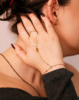 A gold hand chain bracelet with a teardrop-shaped citrine gemstone. The bracelet is connected to a ring that is worn on the index finger. The chain is made of small, delicate links.