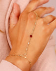 A gold chain bracelet with a red gemstone. The bracelet is designed to wrap around the wrist and finger.