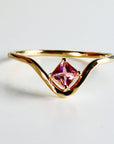 A gold ring with a pink square-shaped gemstone set in the center. The band of the ring is curved and has a V-shape.