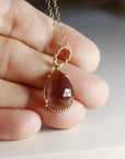 A gold chain necklace with a teardrop-shaped faceted smokey quartz gemstone in a gold prong setting, featuring a twisted rope bail, displayed on a hand.
