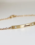 A gold bracelet with a delicate chain and a rectangular bar. The bar is engraved with the name "Emma" and features two small, sparkling gemstones. The bracelet is designed for everyday wear.