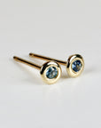 A pair of gold stud earrings with a bezel setting. Each earring features a small, round, light blue sapphire gemstone. The earrings are displayed on a white surface, with the focus on the jewelry.