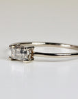 A gold ring with a rectangular, salt and pepper diamond. The diamond is set in a four-prong setting. The band is thin and polished. The ring is displayed on a white surface.