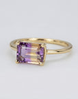 A close-up of a handcrafted ring with a large, rectangular emerald-cut ametrine stone. The gemstone shows a beautiful gradient of deep purple and honey yellow, set in a sleek, minimalist metal setting on a white background.