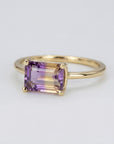 A close-up of a handcrafted ring with a large, rectangular emerald-cut ametrine stone. The gemstone shows a beautiful gradient of deep purple and honey yellow, set in a sleek, minimalist metal setting on a white background.