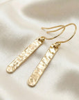 Flat lay of hammered gold filled bar dangle earrings, minimalist gold bar drop earrings on neutral background