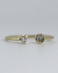 A thin gold open cuff ring with a small, clear diamond in a prong setting on the left and a round, dark iridescent opal on the right.