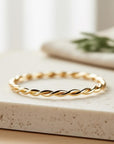 A single gold twisted rope ring rests on a light-colored stone surface, with a blurred green plant in the background.