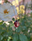 A gold necklace with an oval, faceted, dark pink tourmaline pendant, with a small triangular diamond set below it, hangs in front of a white rose and green foliage.
