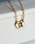 A gold chain necklace with a small heart-shaped pendant engraved with a black Gothic letter "A" rests on a white surface.