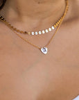 A person wears a layered gold necklace, with a choker made of flat gold discs and a longer chain with a small heart pendant engraved with a white letter "A."