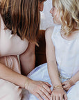 A woman in a light pink top holds the hand of a child in a white dress, both wearing gold cuff bracelets.