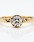 A gold ring with a hammered band and a round, bezel-set diamond is centered on a white reflective surface.