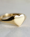 A close-up of a shiny, unengraved gold heart-shaped signet ring on a white surface.