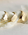 Three gold signet rings of different shapes—a heart, a rectangle, and an oval—lined up on a light-colored, textured fabric.