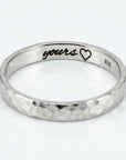 A white gold hammered band ring with "yours" and a heart engraved on the inside.