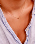 A gold chain necklace with a small bow charm is worn by a person in a light blue shirt, with the charm positioned centrally on the chest.