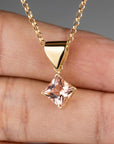 A gold chain necklace with a square, blush-pink gemstone pendant, set in a gold bezel, is worn by a person, visible from the chest up.