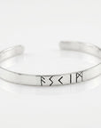 A close-up studio shot of a smooth silver-colored metal cuff bracelet featuring several engraved rune characters on a reflective white surface.