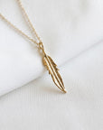 A textured gold feather charm hangs from a delicate chain on an off-white fabric backdrop, viewed at an angle.
