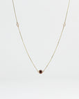 A gold chain necklace with a single red oval gem in the center and two clear round gems spaced evenly on either side, all set in bezels, is displayed against a light gray background.