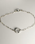 A silver paperclip chain bracelet with a round, flat silver charm featuring a stamped tree design, laid flat on a white surface with the clasp visible.