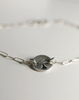A close-up, slightly blurry shot of the round, flat silver charm with a stamped tree design, attached to a silver paperclip chain bracelet on a white surface.