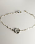 A silver paperclip chain bracelet with a round, flat silver charm featuring a stamped tree design, laid flat on a white surface, showing the full length of the bracelet.
