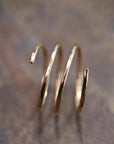 A gold-colored, three-coil spiral ring with hammered texture is shown on a blurred dark brown background.