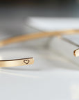 Two gold-colored open cuff bracelets are shown, each with a small engraved heart symbol on one end.