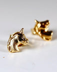 Two gold unicorn head stud earrings resting on a white surface with the foreground earring in sharp focus.