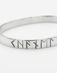 An angled close-up view of a polished silver-colored metal cuff bracelet displaying a sequence of stamped rune symbols.