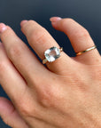 A hand wearing a gold ring with a large cushion-cut light blue gemstone and small flush-set diamonds on the band.