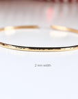 A thin gold hammered open cuff bracelet rests on a white surface with text reading 2 mm width below it.