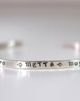 A thin silver cuff bracelet stamped with the word METTA flanked by star symbols against a plain background.