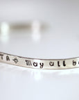 A silver cuff bracelet viewed from an angle, featuring the stamped words May all beings.